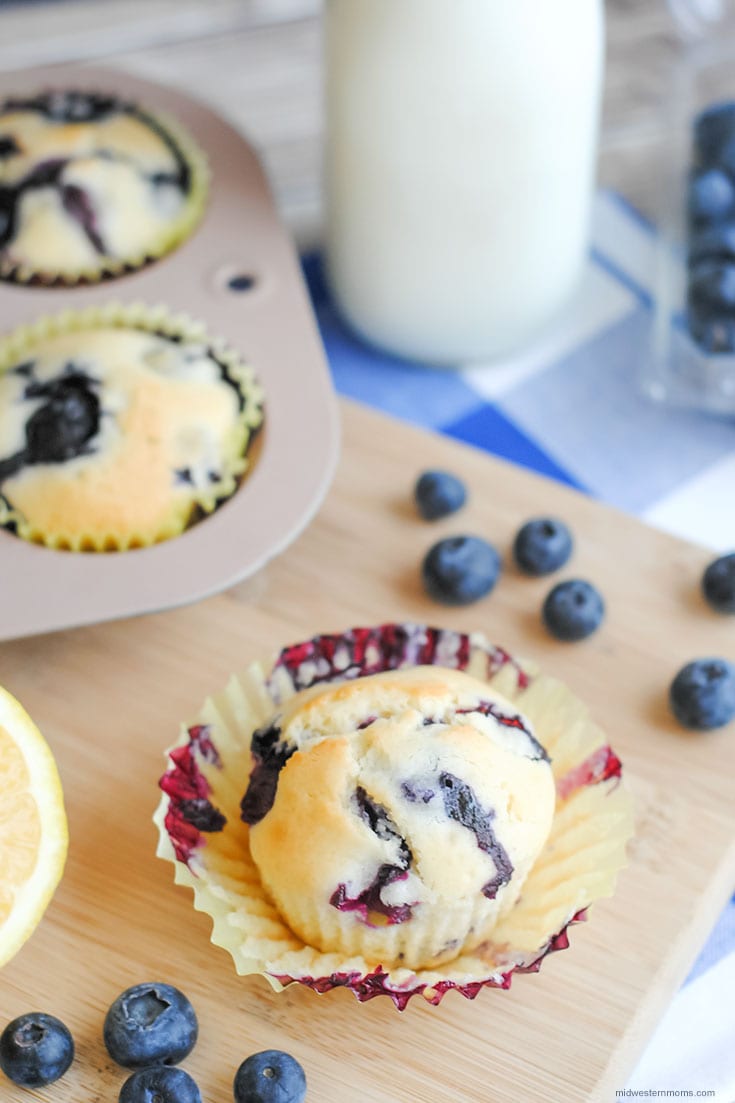 Lemon Blueberry muffin sitting on a wooden cutting board with fresh lemon and blueberries laying around it and a glass of milk.