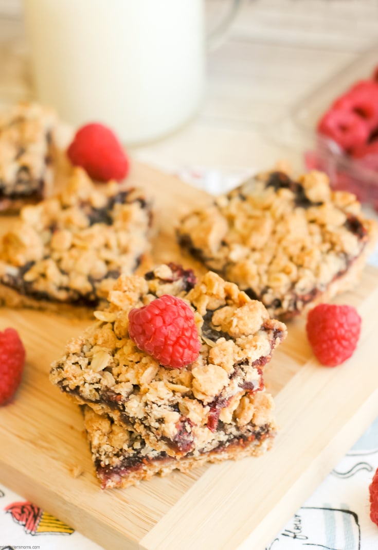 Raspberry Crumble Bars that are topped with a fresh raspberry sitting a wooden cutting board with a glass of milk.