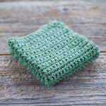 Sage Green crochet dishcloth that is folded in quarters is sitting on old barn wood background. The dishcloth is made with single crochet stitches.