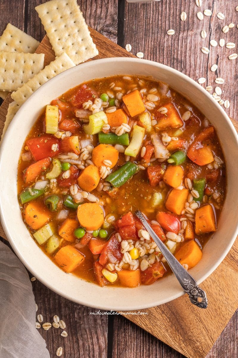 A bowl of hearty vegetable soup that was prepared in a slow cooker on the table with a spoon and crackers.